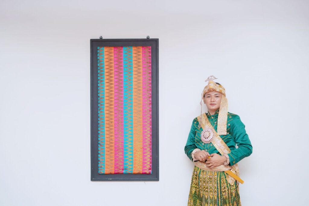 A groom in traditional Makassar attire stands beside a colorful woven tapestry.