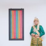 A groom in traditional Makassar attire stands beside a colorful woven tapestry.