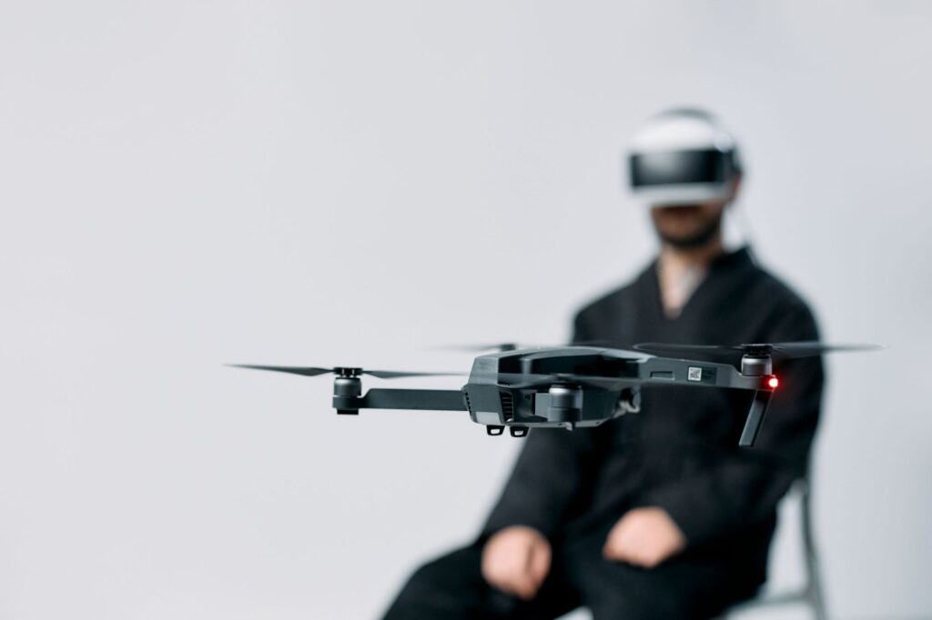 A drone hovers indoors while a person wearing VR goggles holds their hands in anticipation.