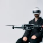 A drone hovers indoors while a person wearing VR goggles holds their hands in anticipation.