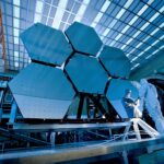 Engineers in protective suits work on telescopic mirrors in a high-tech lab.