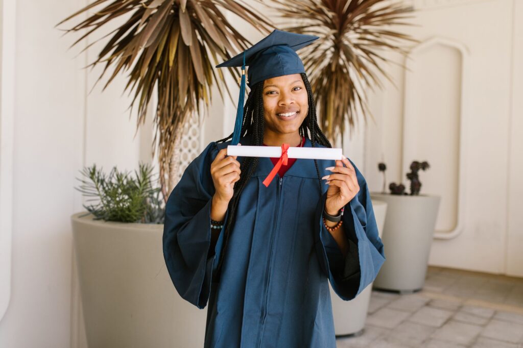 Young woman celebrating graduation with diploma in hand, wearing cap and gown, smiling proudly.
