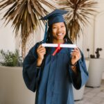 Young woman celebrating graduation with diploma in hand, wearing cap and gown, smiling proudly.