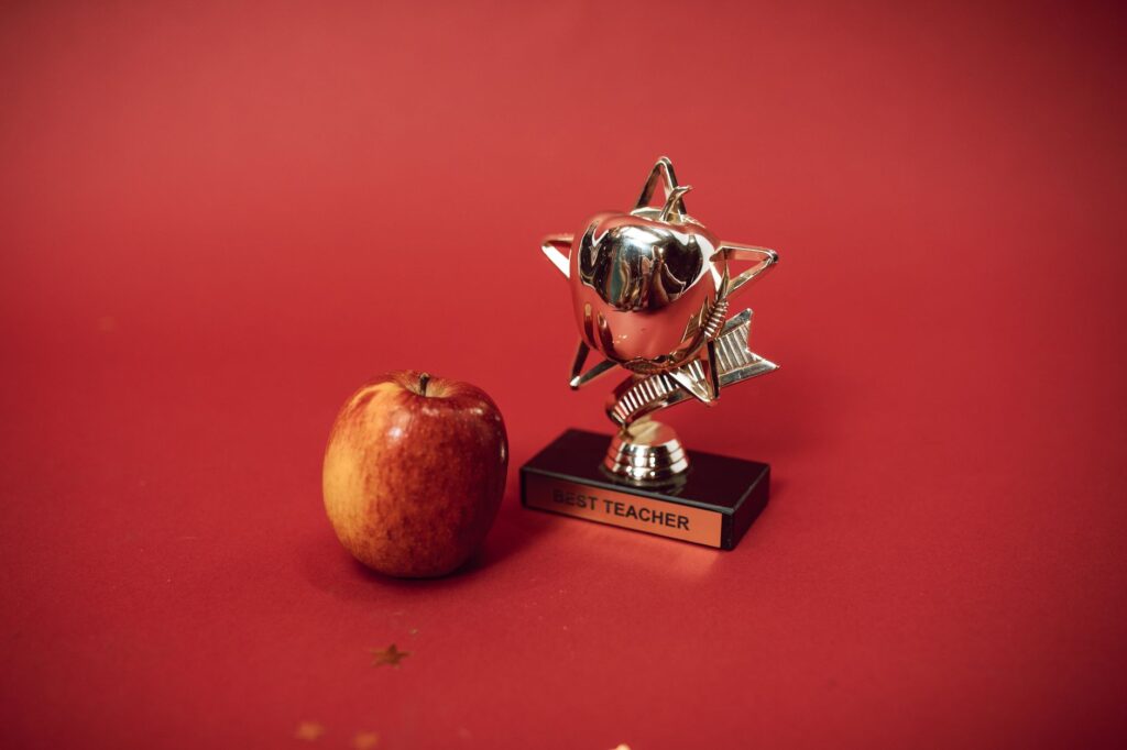 A 'Best Teacher' trophy alongside an apple on a vivid red background symbolizing education and recognition.