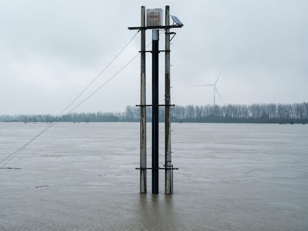 Flooded river landscape featuring a solar panel tower and distant wind turbine under overcast skies.