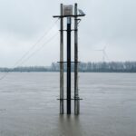 Flooded river landscape featuring a solar panel tower and distant wind turbine under overcast skies.