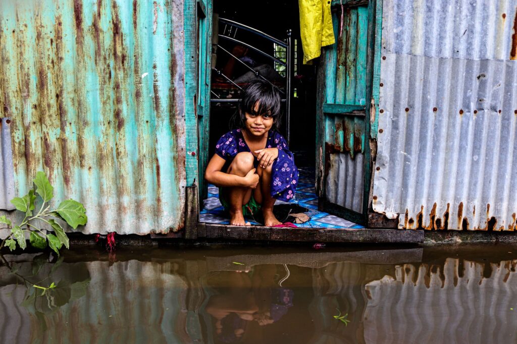A young girl sits in a doorway of a corrugated metal house, surrounded by floodwaters in Bangladesh.