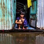 A young girl sits in a doorway of a corrugated metal house, surrounded by floodwaters in Bangladesh.