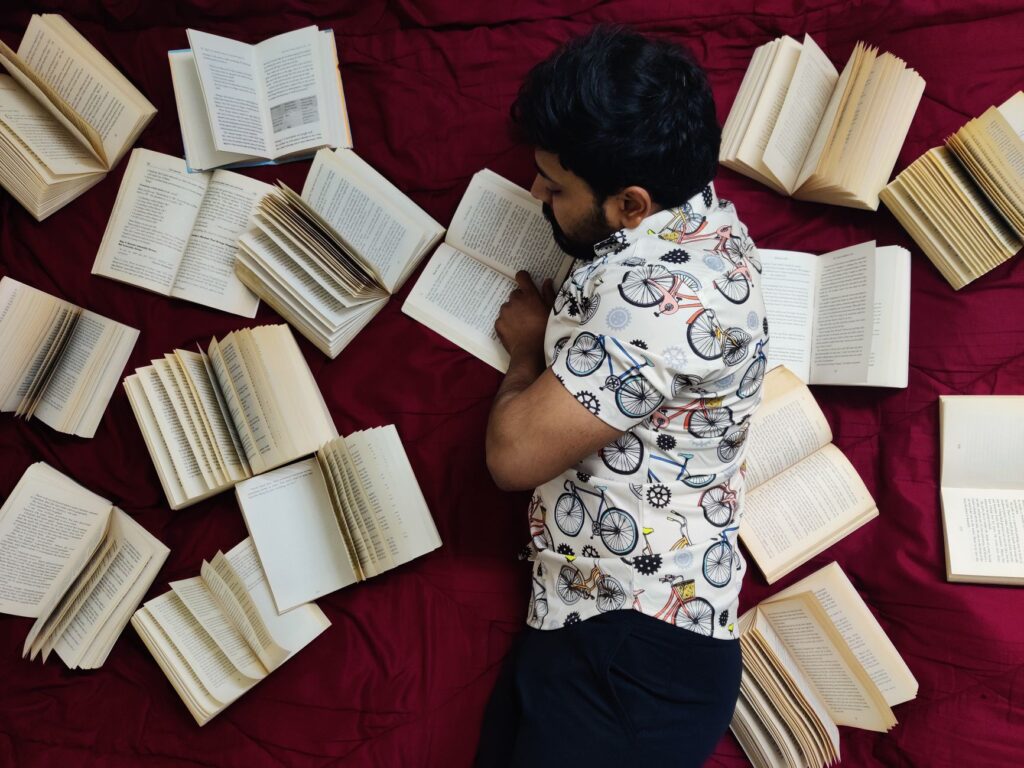 A man lies down reading on a red bedspread surrounded by open books, creating an artistic literary scene.