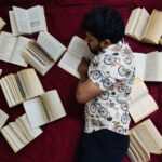 A man lies down reading on a red bedspread surrounded by open books, creating an artistic literary scene.