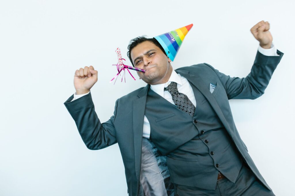 A joyful man in a suit wearing a party hat and blowing a party horn indoors.