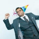 A joyful man in a suit wearing a party hat and blowing a party horn indoors.