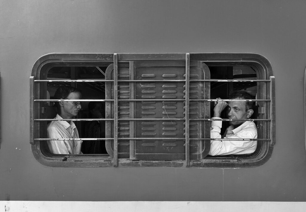 Black and white image of two men sitting inside a train in Bhubaneswar, India, looking out the window.