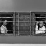 Black and white image of two men sitting inside a train in Bhubaneswar, India, looking out the window.