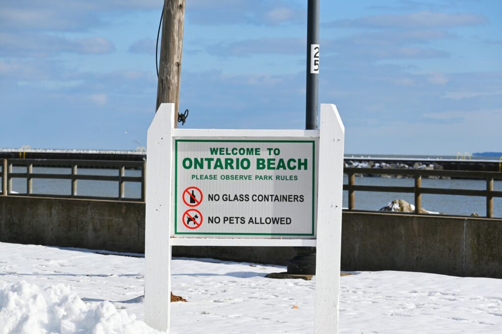 Snowy day at Ontario Beach with pier and welcoming sign, enforcing park rules.