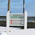 Snowy day at Ontario Beach with pier and welcoming sign, enforcing park rules.