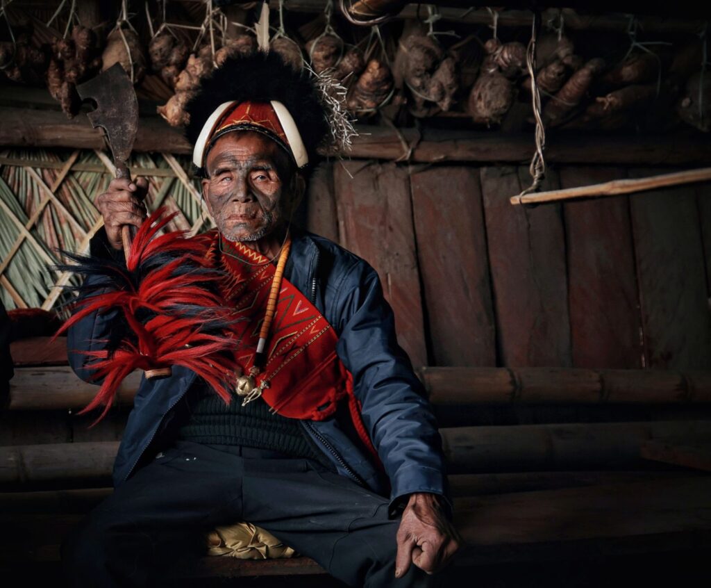 An elderly Naga man wearing tribal attire and holding a machete in a traditional setting.