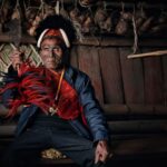 An elderly Naga man wearing tribal attire and holding a machete in a traditional setting.