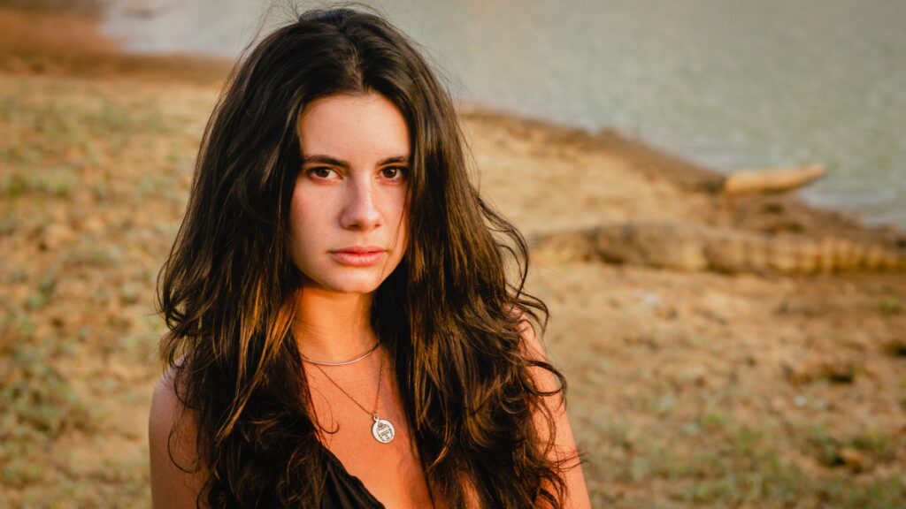 Portrait of a young woman with long hair wearing a silver necklace by a serene lakeside.