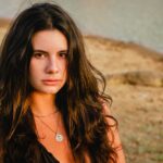Portrait of a young woman with long hair wearing a silver necklace by a serene lakeside.