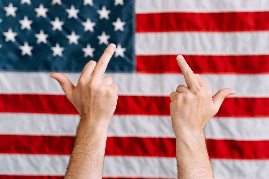 Hands displaying a rebellious gesture in front of the American flag, symbolizing defiance.