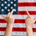 Hands displaying a rebellious gesture in front of the American flag, symbolizing defiance.