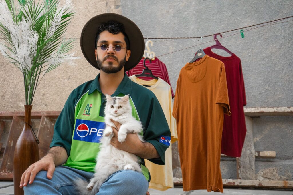 Man in hat with a cat and hanging shirts in a relaxed outdoor setting.