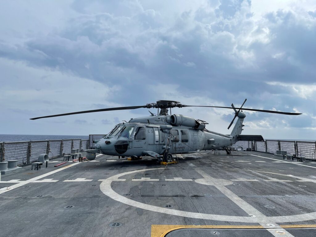 A military helicopter on a naval flight deck under a cloudy sky at sea.