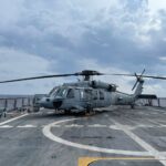 A military helicopter on a naval flight deck under a cloudy sky at sea.