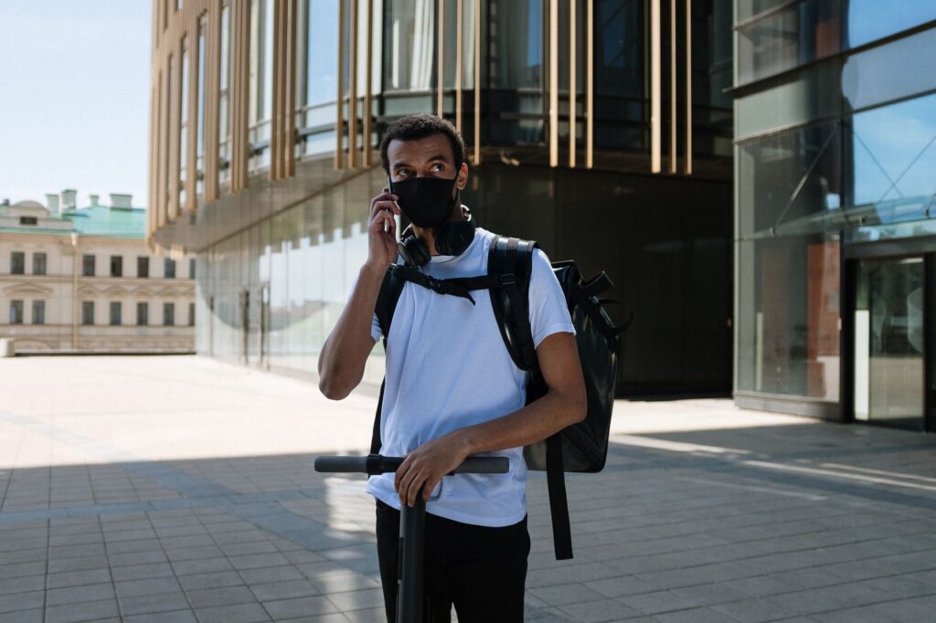 A courier wearing a face mask and riding an electric scooter in a city environment.