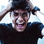 A close-up of a man shouting in frustration, clutching his head with broken eyeglasses on.