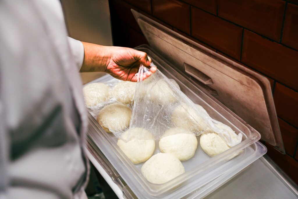 Chef lifting plastic wrap from fresh dough balls in a kitchen setting.
