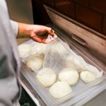 Chef lifting plastic wrap from fresh dough balls in a kitchen setting.