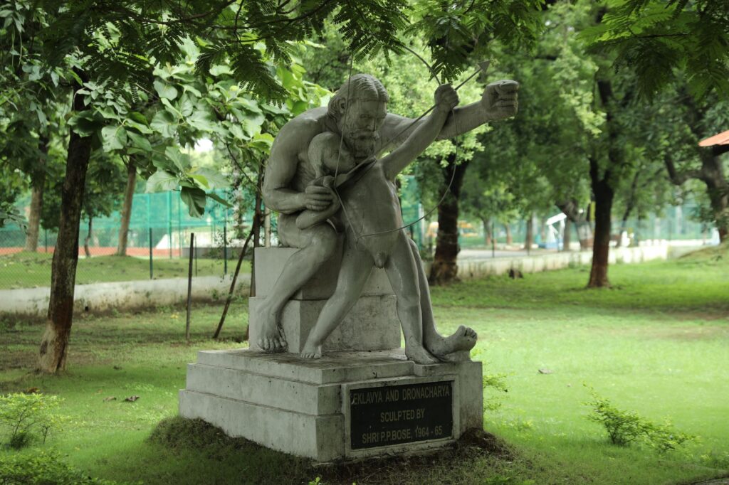Sculpture of Eklavya and Dronacharya, Raipur, India, amidst lush greenery.
