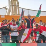 Group of enthusiastic soccer fans holding flags and cheering at a stadium event.