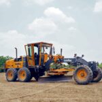 A motor grader working in an open field under a cloudy sky at Sanand, India.