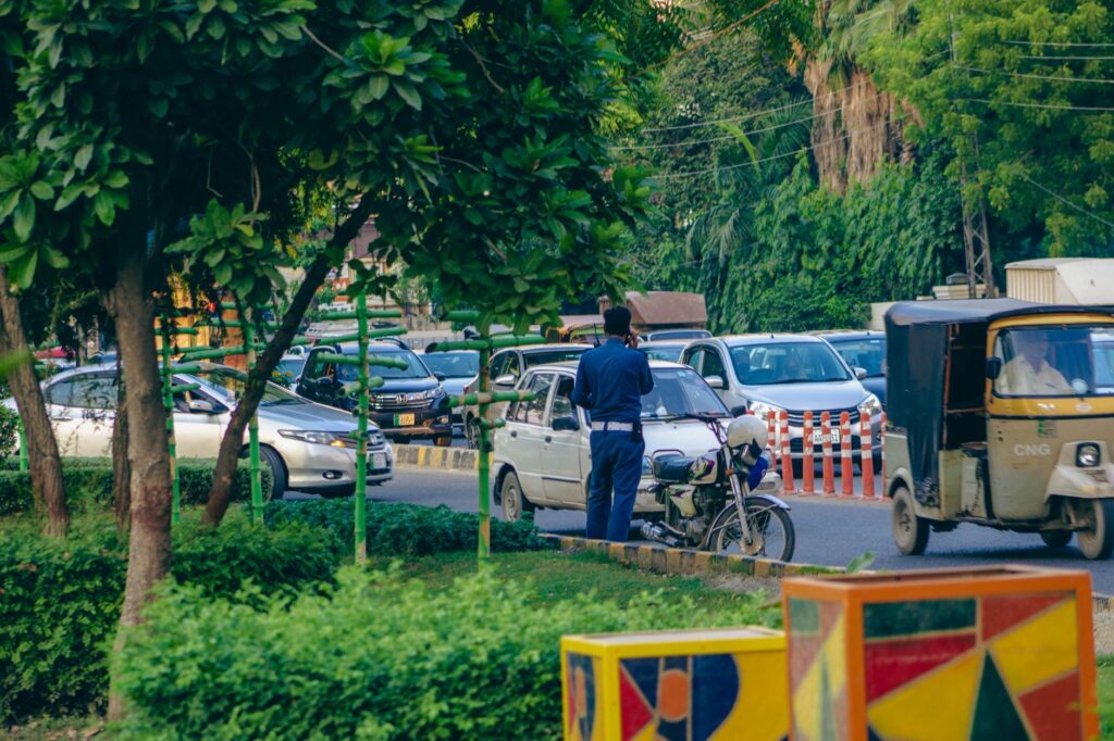A policeman manages heavy traffic on a bustling street in Lahore, Pakistan, amidst greenery and urban chaos.