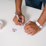 A person wearing a wristband fills out a voting ballot with a pen. Voting stickers visible.