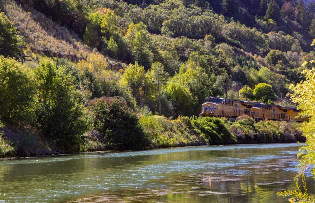 A freight train travels beside a lush, tree-lined river in a hilly landscape.