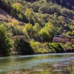 A freight train travels beside a lush, tree-lined river in a hilly landscape.