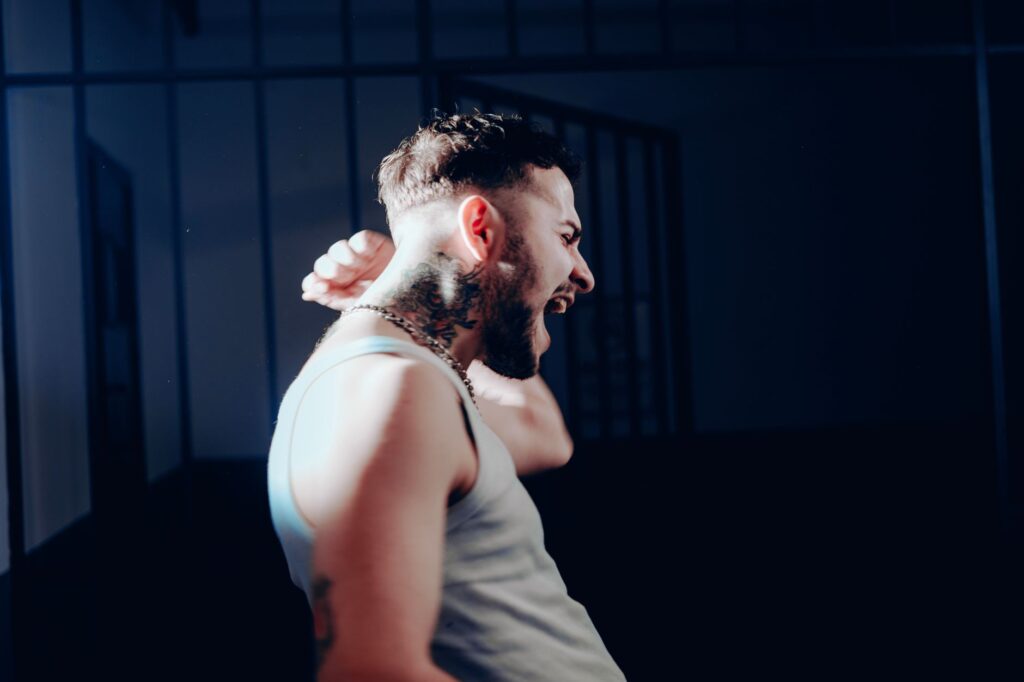 A striking side profile of a man in a tank top, passionately shouting with tattoos visible, set against a dark background.