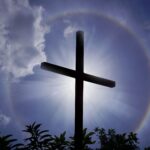 A cross silhouetted against a dramatic sun halo and clouds in India.
