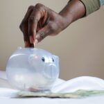 Close-up of a person's hand placing coins into a transparent piggy bank to save money.