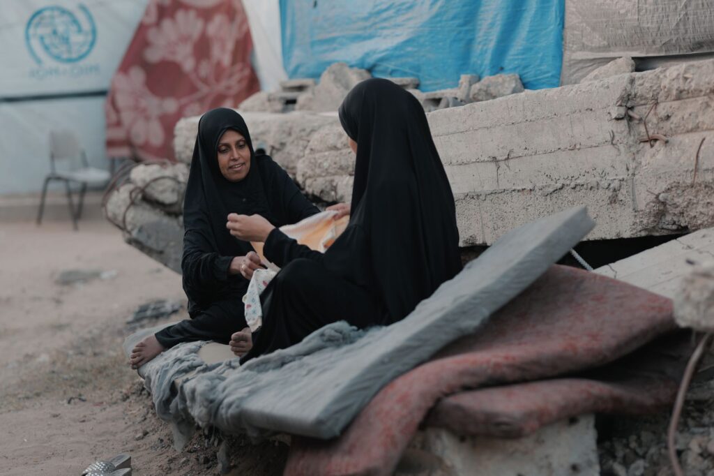 Two women in hijabs sit on rubble in Gaza, sharing a moment of resilience.