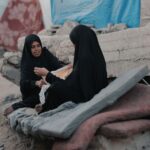 Two women in hijabs sit on rubble in Gaza, sharing a moment of resilience.