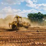 A powerful bulldozer works on a dusty, dirt field under a bright blue sky with cloud cover and trees in the background.