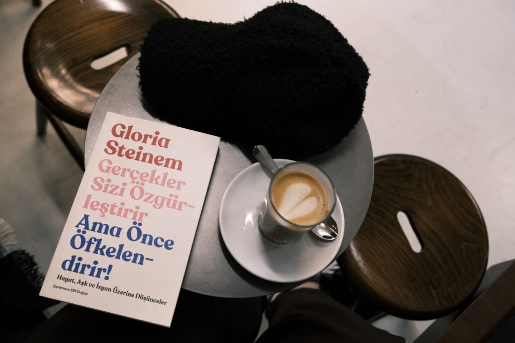 A café setting with a latte, book by Gloria Steinem, and cozy hat on a table.
