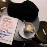 A café setting with a latte, book by Gloria Steinem, and cozy hat on a table.
