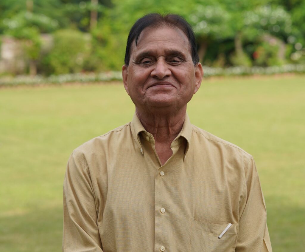 Senior man in a yellow shirt smiling outdoors in a lush green garden.
