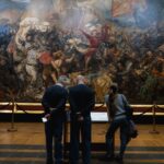 People viewing a large, detailed historical battle painting in a museum setting.